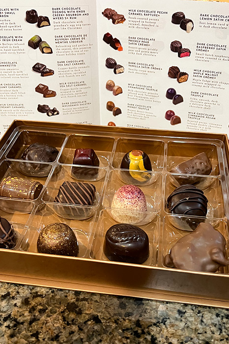 A close-up of a variety box of Ethel M chocolates. In the background, the chocolate guide is on display.