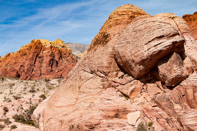 A pyramid-shaped striated red and tan boulder sits in the foreground, while a red and tan mountain  looks smaller in the back against a blue sky with wisps of clouds.