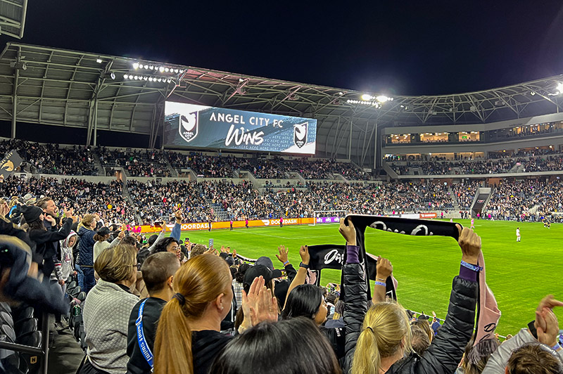 The big screen says "Angel City Wins" at Banc of California stadium. Fans celebrate in the foreground. An ACFC scarf is visible above the heads of the crowd.