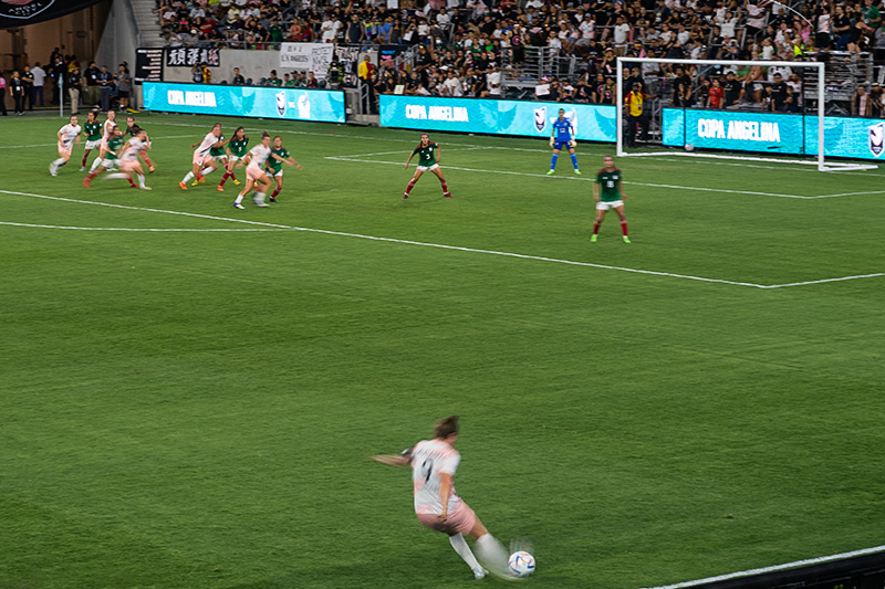 Savannah McCaskill is a blur of motion as she takes a free kick from the sideline. Players from ACFC and the Mexican National team are in blurred motion in the background.