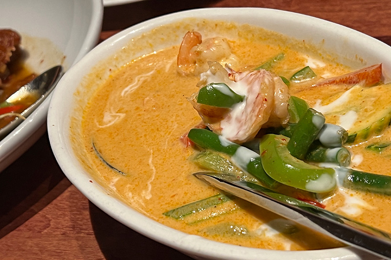 A serving dish of Khao soi sits on a table. The close-up of the dish reveals an orange colored sauce with green peppers and others vegetables peeking out the top.