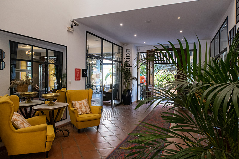 The lobby of the Palacio de Rojas in Valencia, Spain. Two yellow armchairs with yellow decorative pillows sit in front of a mirror at the left. A plant sits in the foreground on the right. The glass wall of reception with "reception" written on the wall beside it sits next to the yellow chairs toward the left, and an open door leads to the patio in the back.