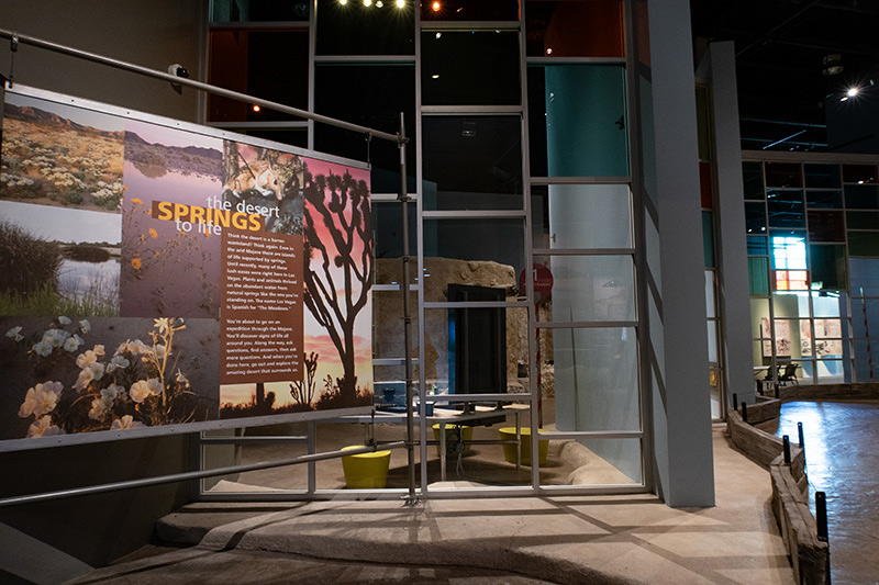 A banner hangs to the left in front of a floor-to-ceiling glass wall. To the right, a path leads into the Origen museum exhibits