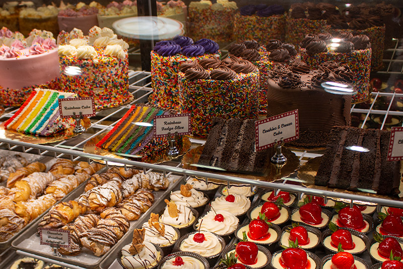 chocolate cake, rainbow colored cake and pastries topped with berries will the display case at Carlo's Bake Shop