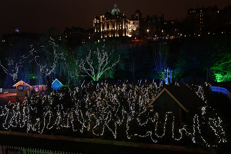 White lights fill the shrubbery and blue and green lighted trees stand behind. Child-sized lighted buildings sit to the left, as behind the historic Bank of Scotland building with its gold-lit exterior and green dome rises on a hill above it.